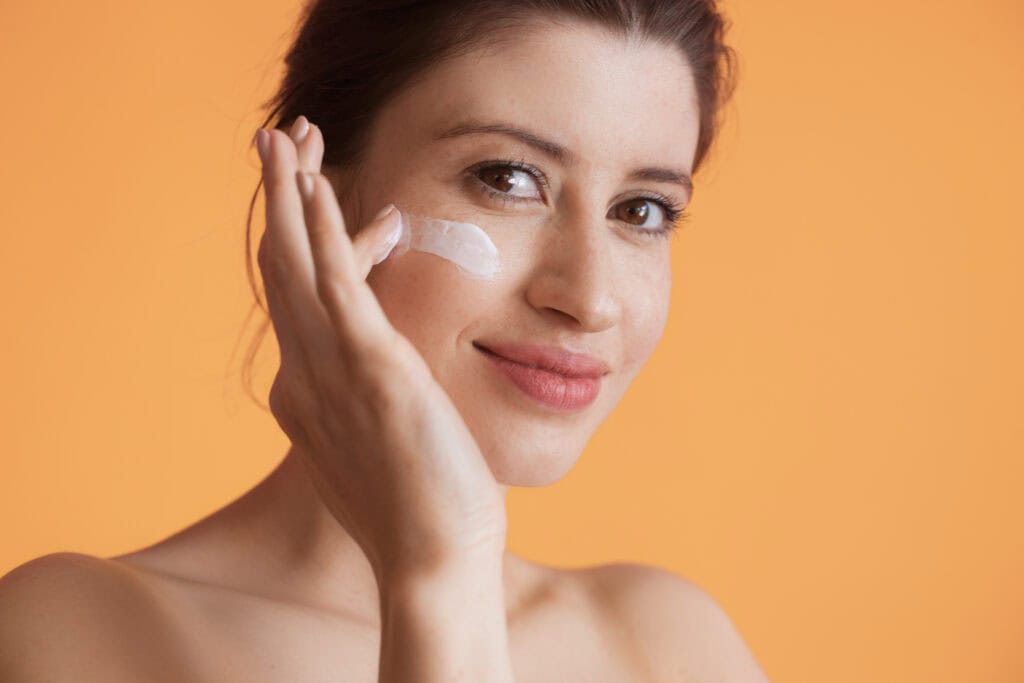 attractive woman with undressed shoulders is posing on a yellow studio wall smiling at camera while applying anti aging cream on her face